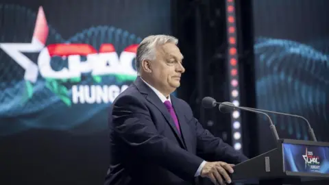 ZOLTAN FISCHER/HUNGARIAN PM HANDOUT Hungary's leader with a blue suit and grey hair stands at a lectern with a flashy white logo behind him