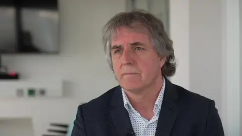 BBC Steve Rotheram, who has grey hair and wears a dark suit, speaks to the camera