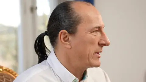 BBC A man in a white shirt with a black pony tail is pictured side-on