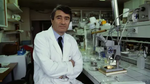 Getty Images Dr Etienne-Emile Baulieu sits in a white lab coat with his arms crossed over his waist inside a lab, with various equipment around him and in the background