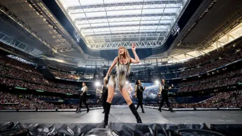 Xavi Torrent/TAS24/Getty Images for TAS Rights Management Taylor Swift stands dancing at front of stage with some backing dancers behind her surrounded by thouands in the crowds at the stadium