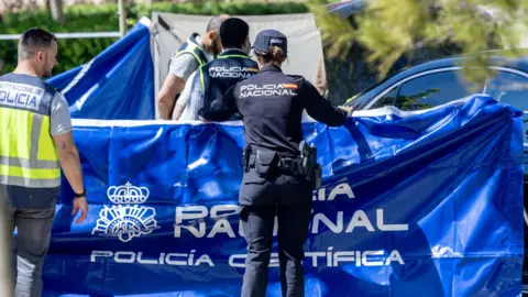 Europa Press via Getty Images Police in Madrid - wearing navy blue uniform that says Policia Nacional - hold up blue tarpaulin to hide the body of Andriy Portnov. You cannot see the body in the photo, but it is clear that this is an active crime scene. It looks to be a sunny day.